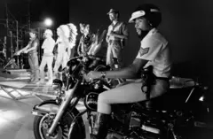 Victor Willis está sentado em uma motocicleta enquanto o restante do grupo Village People se apresenta no palco.