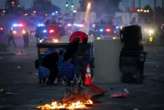 متظاهر يطلق ألعاباً نارية خلال مواجهة بين الشرطة والمتظاهرين بعد اعتقالات متعددة من قبل إدارة الهجرة والجمارك. نيران غير منتشرة مشتعلة، ورجال أمن وسيارات شرطة تملأ المكان.