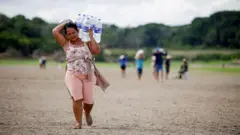 A woman carries large bottles of water across dry ground in Brazil during a heatwave. Other blurred figures in the background are doing the same. 