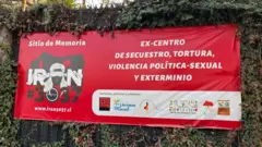 Un primer plano de una pancarta roja situada fuera de los muros del lugar conmemorativo, anteriormente conocido como «Venda Sexy». La pancarta lleva una inscripción en letras blancas y negritas que reza: «antiguo centro de detención, tortura, violencia político-sexual y exterminio». La pancarta también incluye un dibujo de la casa en negro, el nombre «Irán #3037» y el dibujo de una mujer con los ojos vendados.