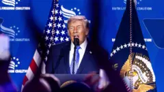 Donald Trump está diante de uma tela azul e de bandeiras