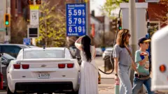 Uma mulher de vestido branco está no centro da imagem. Ela abastece um carro branco em um posto de gasolina em Washington D.C., EUA