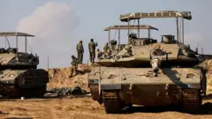 Abasirikare ba Israel bahagaze iruhande rw'ibifaru, mu gihe umwotsi uzamuka inyuma yabo uvuye muri Gaza, hafi y'umupaka wa Gaza, aha bari ni muri Israel, ku itariki ya mbere Ukuboza (12) mu 2023