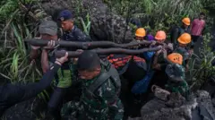 Equipes de resgate vestidas com uniformes de camuflagem e capacetes laranja carregam uma bolsa para cadáveres balanceada em dois grandes postes
