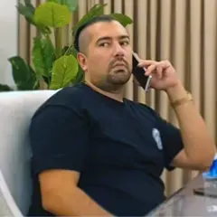 Rawa Majid fala ao telefone sentado em uma cadeira. Ele usa os cabelos penteados para trás, raspados nas laterais, e tem cavanhaque