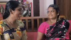 Two women sit on a couch, looking at each other and smiling. They are both wearing colourful patterned Indian clothing, a salwar kameez and a sari. 
