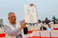 জুলাই সনদ স্বাক্ষরের পর তা ধরে দেখাচ্ছেন অন্তর্বর্তী সরকারের প্রধান উপদেষ্টা মুহাম্মদ ইউনূস, পেছনে বসে আছেন বিভিন্ন রাজনৈতিক দলের নেতারা