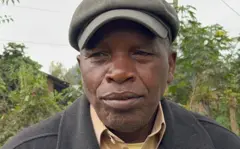 Umugabo wambaye ingofero, ishati n'ikoti, inyuma ye hari ibiti n'inzu