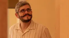 Octávio Santiago sorrindo contidamente em pé, em ambiente interno