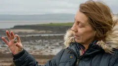 Uma mulher com cabelos castanhos na altura dos ombros, vestindo um casaco azul com capuz de pele. Ela está à direita da imagem e olha para a esquerda, onde segura uma pequena pedra com um fóssil incrustado. Ao fundo, uma paisagem marítima desfocada.