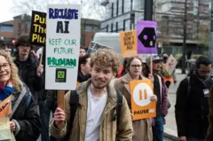 Un joven sonriente sostiene un cartel pidiendo que se regule la IA, para mantener un "futuro humano", en medio de una protesta.