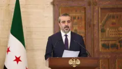 Şeybani gerisinde Suriye bayrağı var bir kürsüde konuşuyor. 