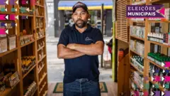 A foto mostra Ednilson, um homemone x bet websitepele morena, vestindo uma camiseta polo azul-marinho, calça jeans e boné. Ele estáone x bet websiteuma loja e olha para a câmeraone x bet websitebraços cruzados.