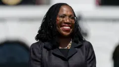 Ketanji Brown Jackson em pé, aparentemente em evento, sorrindo e olhando para o lado