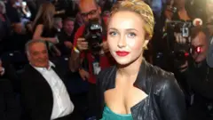Hayden Panettiere, rodeada de fotógrafos, usa o cabelo preso, batom vermelho e um vestido verde com uma jaqueta de couro preta por cima.