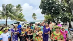 Pobladores de Penrhyn junto al mar sonriendo a la cámara. Las mujeres llevan vestidos coloridos y coronas de flores.