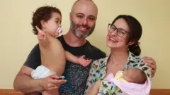 Richard e Rafaela, ele com a filha pequena no colo, sorriem para foto dentronovibet noticiascasa