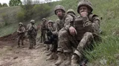 Військові в уніформі сидять на траві, обабіч грунтової дороги. 