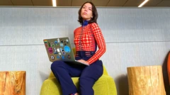 Monica Salley est assise sur un pouf, son ordinateur est placé sur l'une de ses jambes. Elle porte un pantalon violet et un haut manche longe à points rouges munie d'un col violet.