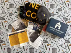 Artículos promocionales y folletos de New Comienzos, incluidos gorros, una bufanda y un libro, colocados sobre un mantel con ilustraciones de utensilios de cocina.