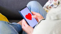 Pessoa sentada olhando para o telefone celular, que mostra um coração na tela.