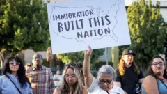 Manifestantes protestam contra uma deportação realizada pelo ICE em Los Angeles, na Califórnia (EUA). Uma manifestante, com a cabeça baixa, exibe um cartaz com os dizeres 'A imigração construiu esta nação'.