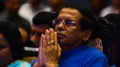 මෛත්‍රීපාල සිරිසේන නිල් පැහැති කමිසයකින් සැරසී සිටී. දෑත් එකට තබා වැඳගෙන සිටින ආකාරයක් පෙන්වයි