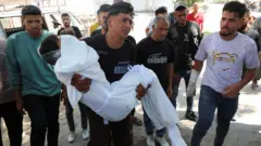 شاب فلسطيني يبكي وهو يحمل بين يديه جثماناً بحجم صغير يبدو لطفل ملفوف بكفن وحوله مجموعة من الرجال.