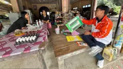 Two women in a shop are talking to Chutiphong Pipoppinyo who is sitting on a bare wooden table and showing them a mockup of a ballot paper. Pipoppinyo is wearing blue jeans, white sneakers and an orange and white jacket. 