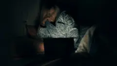 Uma mulher de pijama tem o rosto iluminado por um laptop; ela apoia as mãos no rosto, pensativa.