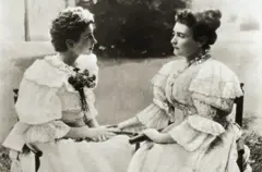 Las dos mujeres, sentadas en un jardín, con vestidos claros de mangas embombadas, con encajes y lazos. Se miran a los ojos y tienen sus manos juntas, pues era su forma de comunicación.