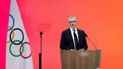 ダークスーツ姿のワッサーマン氏が、五輪マークがついた演台に立ち口を開いている。近くには五輪旗が立てられている