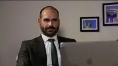 Eduardo Bolsonaro sorrido de forma contida e olhando para lapto
