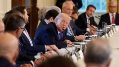 Trump uzun bir masanın ortasında sağında ve solunda resmi giyimli onlarca erkek ve kadın var. Yüzü kameraya dönük, masadakilere bakarak konuşuyor. 