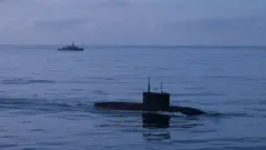 Подводная лодка в надводном положении в море, на заднем плане - военный корабль