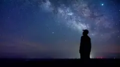 Gece yıldızlı gökyüzüne bakan şapkalı bir adam.