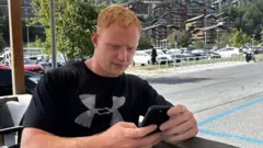 Sol, vestindo uma camiseta preta da UnderArmour, olha para o seu celular. Ele está sentado na esplanada de um café, com um calçadão à beira-mar atrás dele. O tempo está ensolarado.