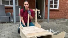 Keira veste camiseta vermelha e luvas de proteção, está agachada atrás do trenó que está fazendo. Ao fundo, aparecem algumas cadeiras de jardim cinza e uma porta aberta que dá para dentro de casa