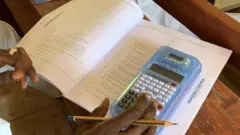 Kalkuleta a kan takardar tambayoyi a kan kujera yayin da hannun wata ɗaliba ke riƙe da fensir