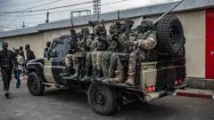 Abarwanyi bo mu mutwe w'inyeshyamba wa M23 bicaye mu modoka ya 'pickup', i Goma, ku itariki ya 6 Gashyantare (2) mu 2026. Bambaye imyenda yijimye, iruhande rwabo hari abandi bantu, n'umwe muri bo na we ufite imbunda arimo kugenda. Abicaye mu modoka bose bafite imbunda. Inyuma hari urukuta rw'inyubako yubakishije isima. 