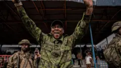 Lt Col Willy Ngoma yambaye imyenda ya gisirikare azamuye amaboko arimo guseka asuhuza abantu. Iruhande rwe hari abasirikare babiri bamurinze. Inyuma hari abandi bantu benshi. Ifoto yo ku itariki ya 6 Gashyantare (2) mu 2025, i Goma, ku kibuga cya Stade de l'Unité.  