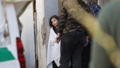 Yüzü yaralı küçük bir kız beyaz bir örtüyle kendini örtmüş, yüzü görünmeyen büyük adamların yanında bir noktaya bakıyor.