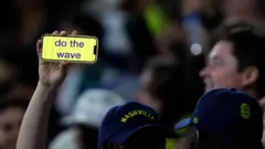 Brazo levantado con la mano sosteniendo un iPhone en cuya pantalla se ve escrito "do the wave" en letras azules y fondo amarillo. De la dueña sólo se ve su cachucha con la palabra Nashville, y la de otra persona a su lado, y en el fondo se ven otras personas borrosas.
