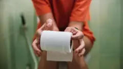 Una mujer sentada en un inodoro con un rollo de papel en los dedos