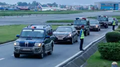 त्रिभुवन विमानस्थलको धावनमार्गनजिकै प्रधानमन्त्री सवार गाडी र उनलाई सुरक्षा दिन खटेको टोलीका सवारीसाधनहरू, बाटोको छेउमा दुई प्रहरी उभिएका छन्