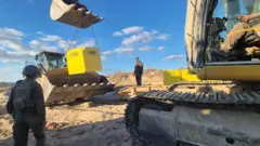 Las tropas israelíes observan cómo una excavadora coloca un bloque amarillo en su sitio. El trabajo cuenta con el apoyo de otro vehículo de construcción, y el bloque queda suspendido en el aire mediante una cadena fijada a la excavadora.