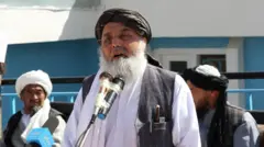 عبدالله سرحدی، والی قبلی بامیان با پیراهن سفید و لنگی و واسکت سیاه در برابر دو مایک صحبت می‌کند، دو فرد دیگر در پشت سر او نشسته‌اند.