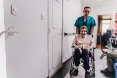 Senhora sendo empurrada em cadeira de rodas por profissional, com outros idosos ao fundo
