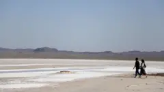 Homem e mulher andam ao longo do leito seco do lago salgado de Hoz-e Soltan em Qom, ao sul de Teerã. Pode-se observar o sal na superfície e as montanhas à distância