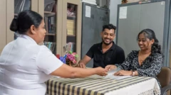 සුදු පැහැති නිල ඇඳුමකින් සැරසී සිටින පවුල් සෞඛ්‍ය සේවා නිලධාරිනියකගේ මේසය ඉදිරිපස අසුන්ගෙන සිටින සිනාමුසු මුහුණින් යුතු තරුණ යුවළක් 